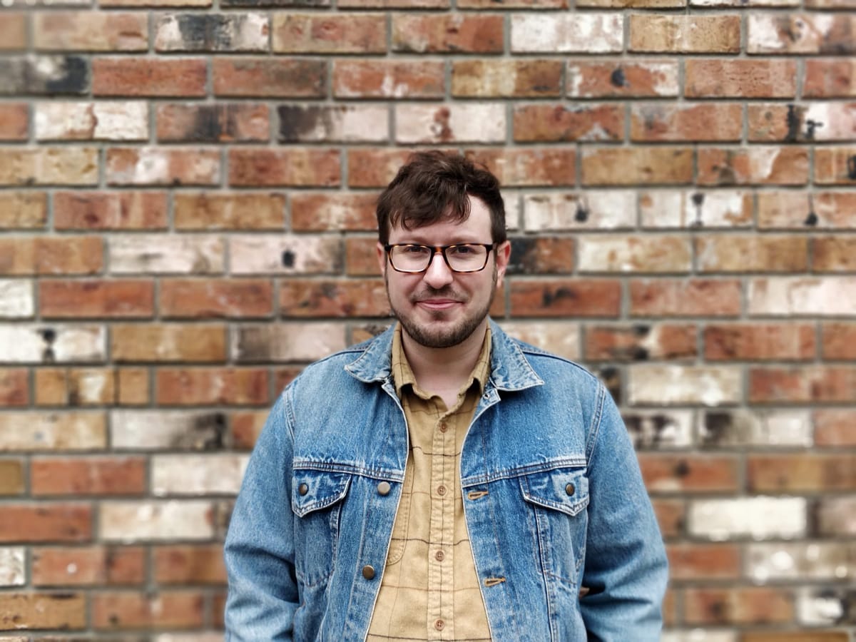 Picture of Phil Taylor-Parker, founder of Intentionally Orange, in a beige flannel shirt and jean jacket, against a brick wall backdrop.